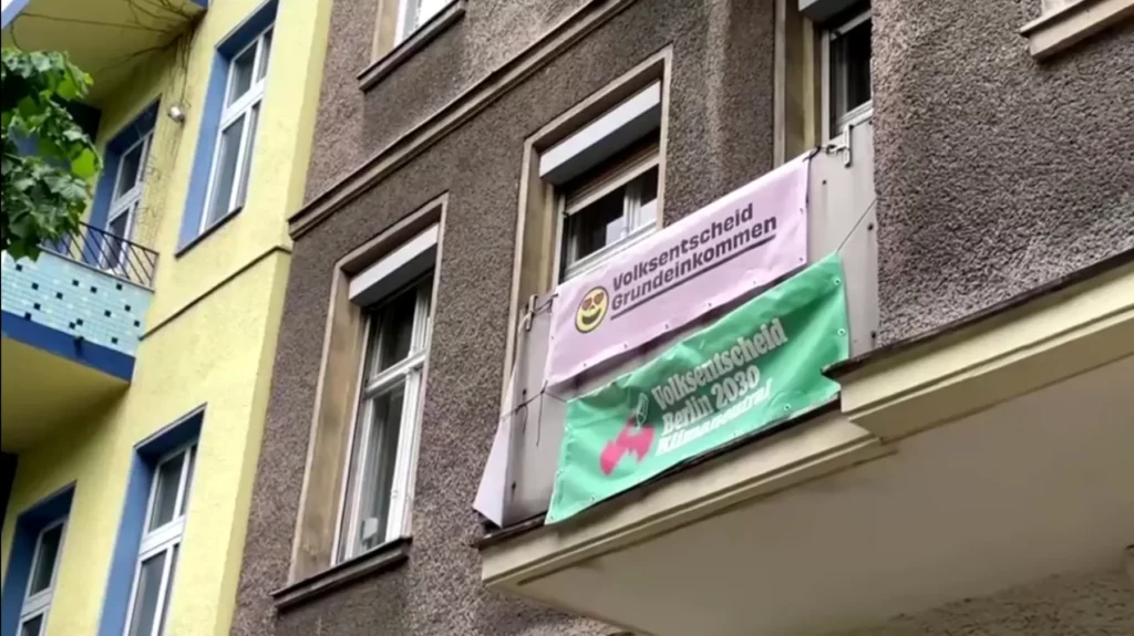 Screenshot von YouTube Video. Hauswand mit 2 Bannern. Smiley mit Herzaugen Volksentscheid Grundeinkommen. Und Banner Volksentscheid Berlin 2030 Klimaneutral
