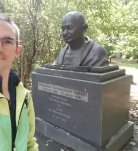 Mohandas Karamchand Gandhi rainerroessler natur im freien gedenkort statue deutschland hannover 2025 Oberkörper Abbild von Mohandas Karamchand Gandhi auf einem dunklen Fundament. Auf dem Fundament steht auf der Vorderseite: Mohandas Karamchand Gandhi 2. Oktober 1869 - 30. Januar 1948 Enthüllt durch THE Prime Minister of India Shri Narendra Modi 12. April 2015 Die Persönlichkeit rainerroessler.de steht davor und schaut in die Kamera. Ist zur Hälfte zu sehen. Mit Brille. Es ist sonnig und eine grüne Landschaft ist zu sehen.